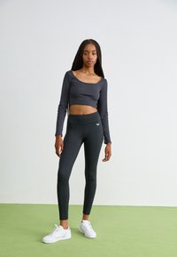 Černý zkrácený dlouhý rukáv top a tmavě šedé legíny z elastické tkaniny. Model má na sobě bílé tenisky. Jednobarevné, minimalistický design.