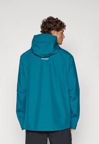Teal waterproof jacket with a hood, featuring a relaxed fit, raglan sleeves, and the "Mammut" logo on the back. Smooth texture.