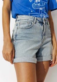 Personne portant un short en denim léger bleu roulé et un t-shirt graphique bleu, montré de mi-cuisse à taille.