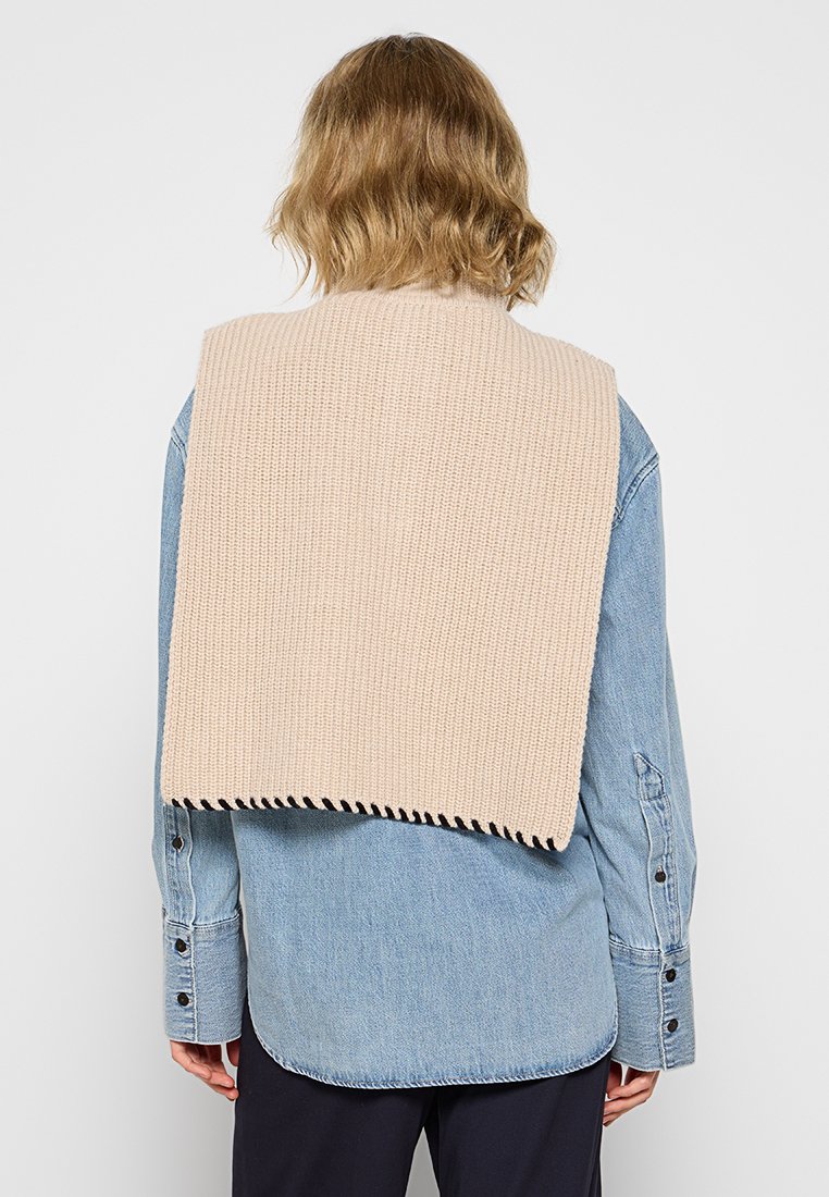 Beige Strickpullover, der über ein blaues Jeanshemd gelegt ist, mit einem gewellten Abschluss und einer strukturierten Oberfläche. Sichtbare Knöpfe am Hemd.