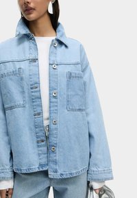 Veste en denim bleu clair avec un col, deux poches avant et des boutons-pression argentés. Coupe oversize avec des détails de couture visibles.