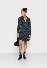 Vestido preto com flores, mangas longas, decote em V e barra larga. Apresenta padrões florais coloridos. Combinado com uma bolsa preta e botas de tornozelo.