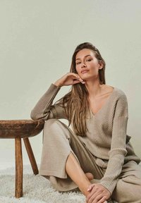 Beige gestreifter Strickpullover mit V-Ausschnitt und langen Ärmeln, kombiniert mit passenden weit geschnittenen Hosen. Rustikaler Holztisch in der Nähe und strukturierter Teppich.