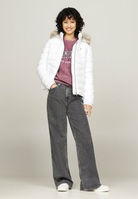 Giacca a vento bianca con cappuccio in pelliccia sintetica, abbinata a una felpa viola e jeans grigi a gamba larga. Sneakers bianche completano l'outfit.