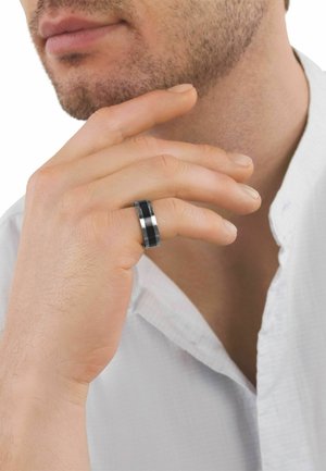 Mann mit leichtem Bartschatten, der ein weißes Hemd trägt, sein Kinn berührt und einen schwarzen und silbernen Ring am rechten Mittelfinger trägt.