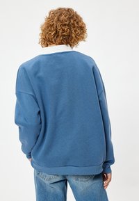 Blauwe sweatshirt met een witte kraag, losse pasvorm, zachte textuur, geribde boorden en zoom. Bevat minimale details op de achterkant.
