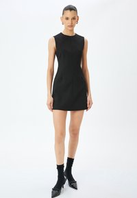 Mini abito nero senza maniche con una silhouette aderente, caratterizzato da un colletto rotondo e dettagli di cucitura frontale, abbinato a calze nere e scarpe con punta.