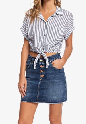Femme portant une chemise nouée à manches courtes à rayures verticales et une jupe en denim bleue avec boutons sur le devant et poches.