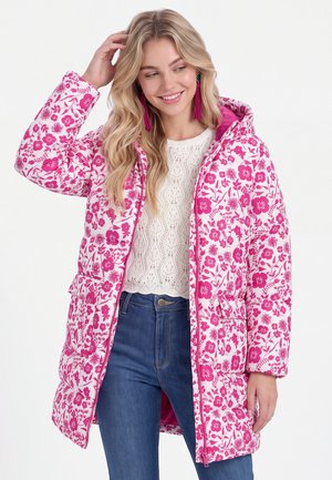Frau trägt einen weißen Strickpullover und blaue Jeans mit einer weiß-rosa floral gemusterten Kapuzen-Steppjacke, lächelt und berührt ihr Haar.