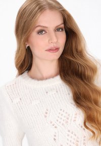 Biała dziana sweter z teksturowanym wzorem i okrągłym dekoltem. Długie, faliste włosy spływają na jedno ramię. Delikatny makijaż i kolczyki.