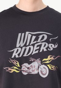 Black cotton t-shirt featuring the text "WILD RIDERS" in silver and a motorcycle graphic with flames in red, orange, and yellow.