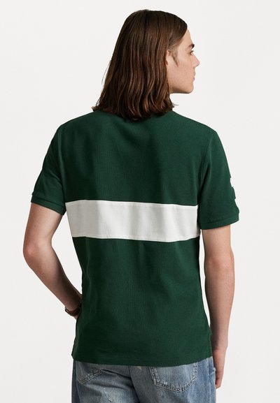 Zaļš kokvilnas T-krekls ar baltu horizontālu svītru aizmugurē, īsām piedurknēm un brīvu piegriezumu. Vienkāršs dizains, bez redzamām logo.