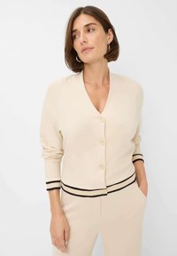 Vrouw in een crèmekleurige cardigan met knopen en zwarte afwerking op de mouwen en zoom, gecombineerd met bijpassende crèmekleurige broek, staand tegen een witte achtergrond.