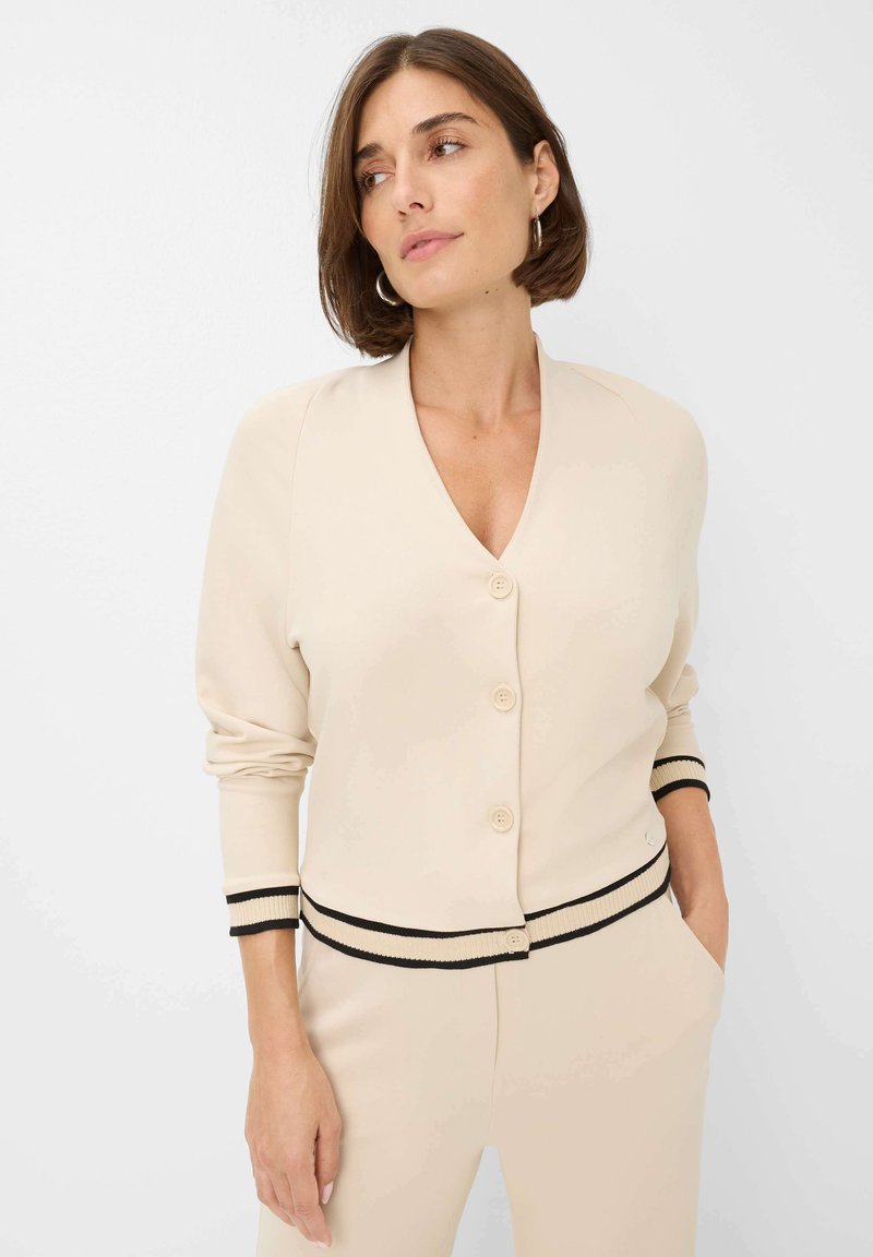 Vrouw in een crèmekleurige cardigan met knopen en zwarte afwerking op de mouwen en zoom, gecombineerd met bijpassende crèmekleurige broek, staand tegen een witte achtergrond.
