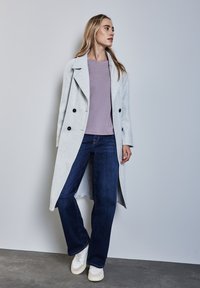 Helles graues langes Coat mit schwarzen Knöpfen, kombiniert mit einem lila Oberteil und dunkelblauen weiten Jeans. Weiße Sneakers mit strukturierten Akzenten.