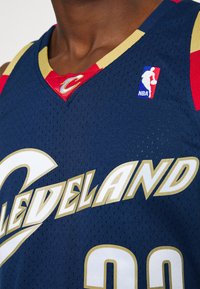 Maglia da basket blu navy con accenti rossi e rifiniture dorate. Realizzata in tessuto a rete traspirante, presenta un grande testo "CLEVELAND" e una toppa del logo NBA sulla spalla.