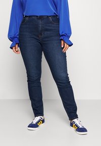 Jeans ajustados de cintura alta azul oscuro con cinco bolsillos, combinados con una blusa azul de manga larga con mangas abullonadas y zapatillas deportivas blancas.
