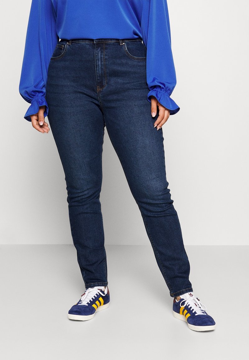 Jeans ajustados de cintura alta azul oscuro con cinco bolsillos, combinados con una blusa azul de manga larga con mangas abullonadas y zapatillas deportivas blancas.