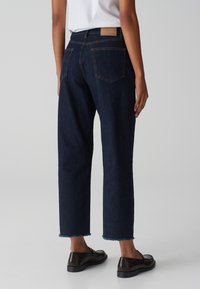 Jeans in denim blu scuro con taglio dritto, orlo sfrangiato e cuciture a contrasto. Presentano due tasche posteriori e una toppa in pelle marrone.