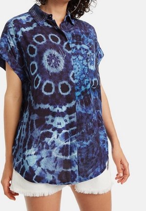 Frau trägt ein lockeres, kurzärmeliges Hemd mit Knöpfen und blau-schwarzen Batikmustern, kombiniert mit weißen, ausgefransten Shorts.