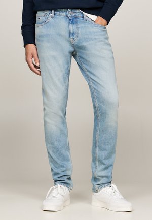 Light blue denim jeans with a slim fit, five-pocket design, subtle fade, and branded patch on the waistband, paired with white sneakers.