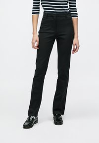 Pantalon noir sur mesure avec une coupe droite, doté de poches avant et d'une finition en tissu lisse. Le modèle porte des mocassins noirs.