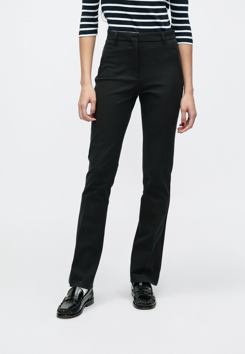Pantalon noir sur mesure avec une coupe droite, doté de poches avant et d'une finition en tissu lisse. Le modèle porte des mocassins noirs.