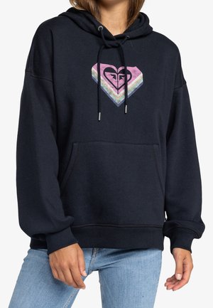 Personne portant un sweat à capuche noir avec un logo cœur aux couleurs de l'arc-en-ciel sur la poitrine et un jean bleu clair.