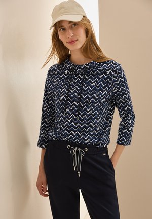 Marineblauw en witte zigzagpatroon top met een ronde halslijn en driekwart mouwen, gecombineerd met zwarte jogpants met trekkoord en een beige pet.