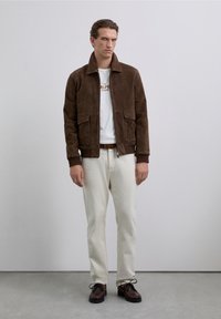 Man wearing brown suede jacket, white graphic t-shirt, cream pants, brown belt, and dark brown shoes standing against a plain gray wall.