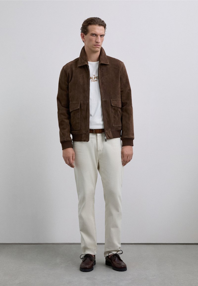 Uomo in giacca scamosciata marrone, maglietta bianca con grafica, pantaloni crema, cintura marrone e scarpe marroni scure, in piedi contro un muro grigio semplice.