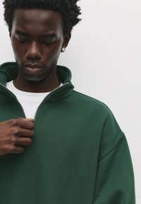 Grøn lynlåsdunket sweatshirt med høj krave, blød tekstur og en hvid t-shirt under, der har subtile syningsdetaljer.