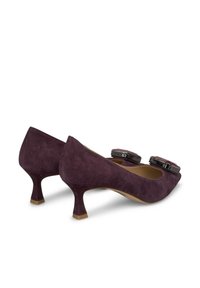 Talons en suede violet avec un bout rond, un petit talon kitten et de grands accents décoratifs sur le devant. Texture lisse et léger éclat.
