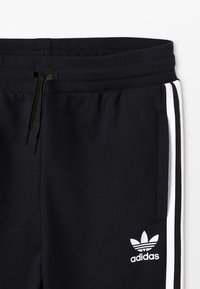 Černé tepláky s páskem na stažení v pase, bílými bočními pruhy a bílým logem Adidas. Materiál vypadá měkký a texturovaný.