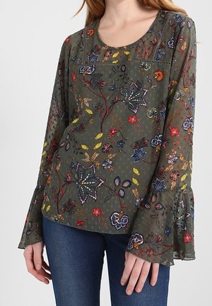 Femme portant un chemisier transparent vert olive avec une broderie florale colorée et des manches évasées, associé à un jean bleu foncé.