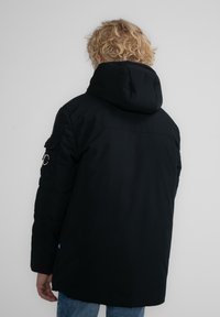 Svart pufferjacka med huva, försedd med ett märke på vänster ärm och en slät, matt yta. Rak skärning med sidofickor.
