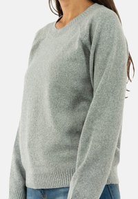 Svetlo siva jopica z mehko teksturo, okrogli ovratnik in rebrasti manšeti. Ima subtilen sijaj, oprijet kroj in dolge rokave.