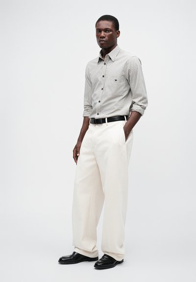 Stripet skjorte i grått og hvitt, parret med vide kremfargede bukser. Svart belte, glinsende svarte sko og oppbrettede ermer gir kontrasterende detaljer.