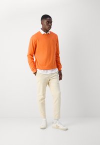 Oranssi huppari pyöreällä kaula-aukolla, valkoinen kauluspaita alla, beige chinot ja valkoiset lenkkitossut beigeillä yksityiskohdilla.