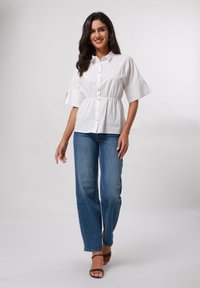 Camicia bianca con bottoni, vita arricciata e maniche ampie, abbinata a jeans blu a gamba dritta e sandali neri con cinturini.