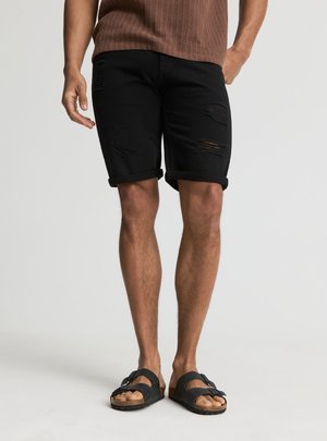 Mężczyzna ubrany w czarne, podarte szorty z podwiniętym dołem, brązowy bezrękawnik i czarne sandały na klamry, stojący na jednolitym tle.