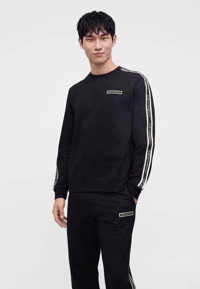 Млад мъж, облечен в черен спортен екип EA7 Emporio Armani с брандирани ивици на ръкавите и панталоните, стои пред обикновен светъл фон.