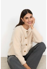 Beige fleecekofta med guldknappar, rund halsringning och två fickor. Modellen har på sig grå byxor och sitter framför en neutral bakgrund.