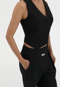 Black pinstriped vest with a deep V-neck, front buttons, and cropped design, paired with high-waisted pants featuring side pockets and logo patch.