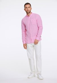 Camicia rosa a maniche lunghe con bottoni, realizzata in un tessuto testurizzato e dotata di taschino, abbinata a pantaloni bianchi e sneakers bianche.