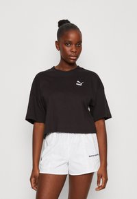 Maglietta nera cropped con scollatura rotonda e maniche corte, caratterizzata da un logo Puma bianco sul petto. Abbinata a pantaloni corti bianchi.