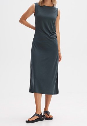 Ärmelloses, dunkelgrünes Midikleid aus glattem Stoff mit rundem Ausschnitt, seitlichem Drapée-Detail und Seitenschlitzen. Kombiniert mit schwarzen flachen Sandalen.