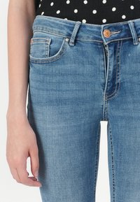 Tejanos de mezclilla azul claro con un ligero desgaste, diseño clásico de cinco bolsillos y cierre de botón. Presenta un fino pespunte visible a lo largo de las costuras.