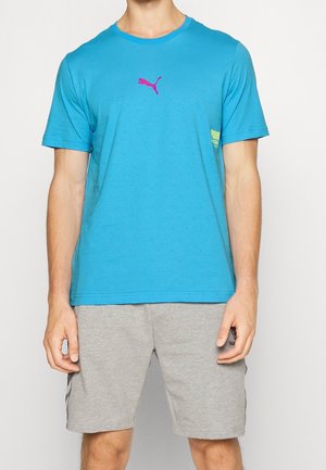 Mężczyzna w niebieskiej koszulce Puma z różowym logo i jasnoszortach stoi na jednolitym tle.