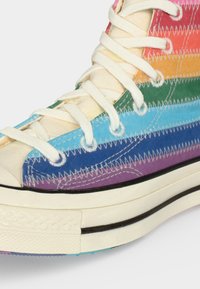Υψηλό sneaker με λευκά κορδόνια και επάνω μέρος από καμβά με ρίγες στο χρώμα του ουράνιου τόξου, με ορατή λευκή ζιγκ-ζαγκ ραφή και χοντρή λευκή καουτσούκ σόλα.